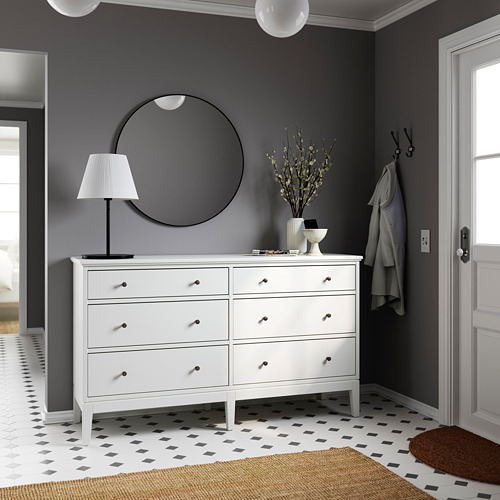 Dormitorio moderno gris con tocador blanco, espejo redondo, lámpara y jarrón floral.