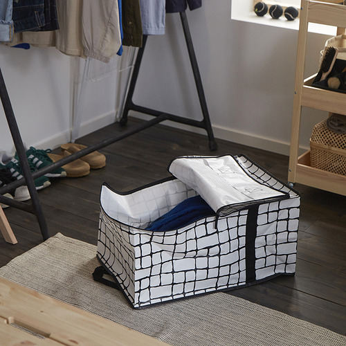 Una bolsa de almacenamiento GÖRSNYGG sobre suelo de madera, con patrón de cuadrícula gris-negro, parcialmente llena de ropa, cerca de un zapatero.