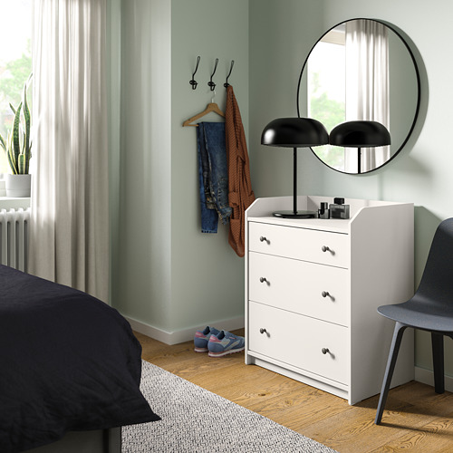Escena del dormitorio: baúl HAUGA blanco, espejo redondo, lámpara negra, silla gris, cama oscura, suelo de madera.