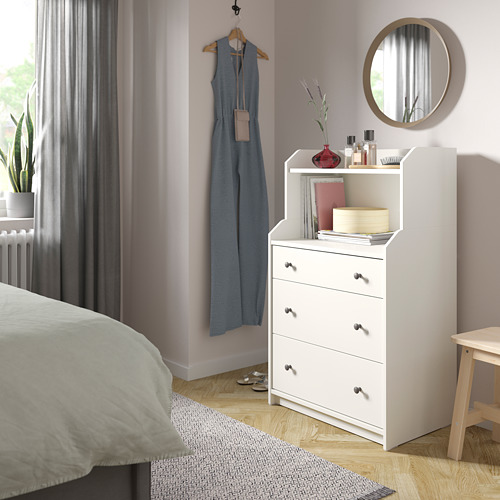 Acogedora habitación con arcón HAUGA blanco, suelo de madera clara, cortinas grises y plantas.