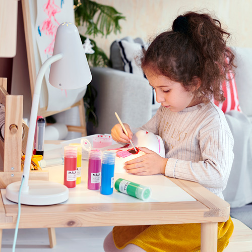Niña pintando en la mesa con colores, con suéter gris y pantalón amarillo.
