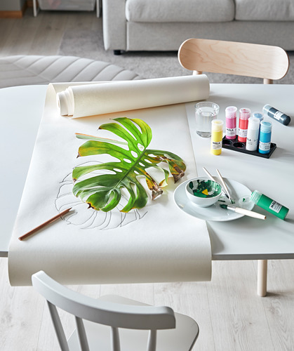 Un juego de mesa de manualidades con pinturas MÅLA, una gran hoja verde, pinceles y un rollo de papel. Diseño minimalista con tonos blancos y madera.