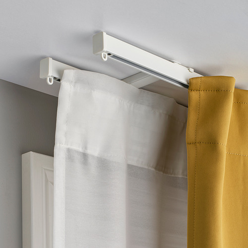 Rieles de cortina blancos montados en el techo con dos capas de cortinas. Las cortinas interiores blancas transparentes y exteriores mostaza cuelgan suavemente de los planeadores.