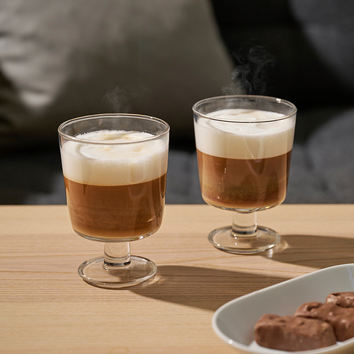 Dos bebidas de café humeantes en tazas anchas de vidrio sobre una mesa de madera. A su lado, un cuenco de golosinas de chocolate.