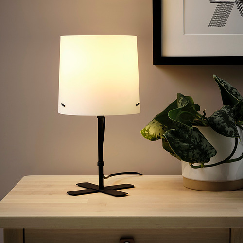 Una pequeña y moderna lámpara sobre una mesa de madera. La pantalla está iluminada, proporcionando una luz suave y sin deslumbramiento, mejorando el ambiente de la habitación. A su lado hay una planta.