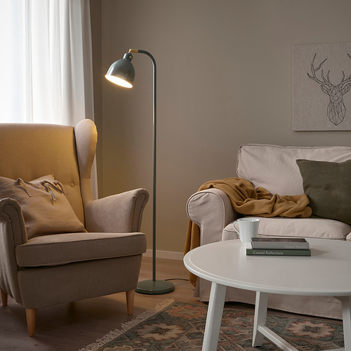 Acogedora sala de estar con lámpara RÖDFLIK, silla beige, sofá, manta, libros y alfombra. La lámpara se mantiene alta cerca de la ventana, proyectando una luz cálida.