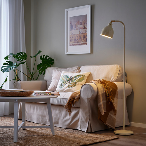 Acogedora sala de estar con lámpara RÖDFLIK, sofá beige, almohadas, arte del paisaje urbano, manta, mesa de café y planta de monstera.
