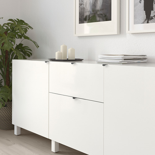 Mueble de almacenaje blanco con manijas modernas, sosteniendo velas y libros contra una pared con fotos enmarcadas y una planta.