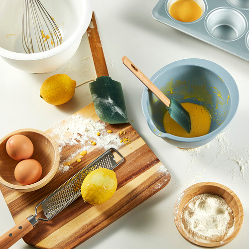 Una escena de cocina con ingredientes y herramientas para hornear, incluido un tazón con pico, batidor, espátula y rallador, listo para mezclar y preparar alimentos.