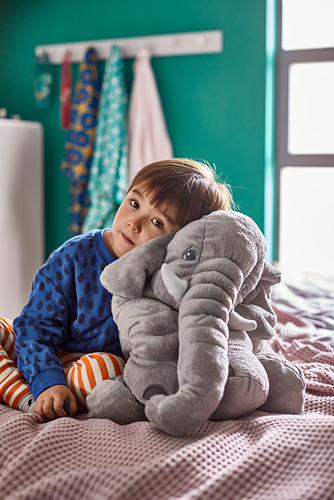 Niño joven en la cama sosteniendo un gran juguete de peluche de elefante JÄTTESTOR gris. Niño en pijama azul, juguete con tronco suave y orejas caídas.