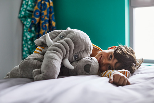 Un niño está acostado en una cama, abrazando un gran elefante gris de peluche.