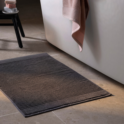 Una alfombra de baño gris oscuro llamada ALSTERN en el suelo junto a una bañera. Está hecho de algodón puro, rectangular y con textura acanalada.