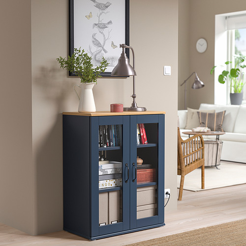 Gabinete SKRUVBY azul con puertas de vidrio, manijas rústicas y planta cerca.
