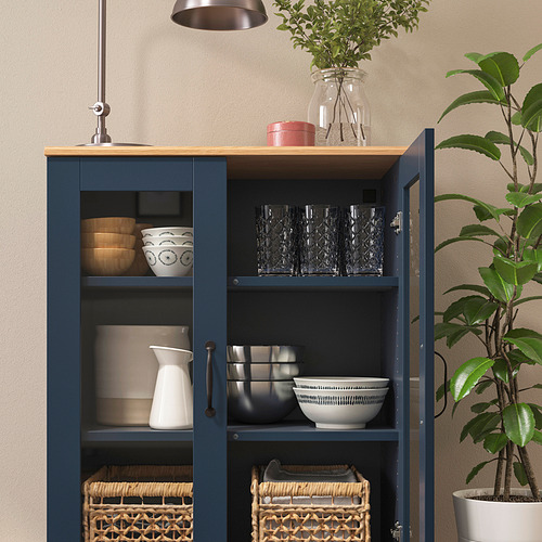 Gabinete azul con puertas de vidrio que exhiben tazones, vasos y cestas; Tapa de roble, vegetación al lado.