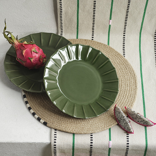 El plato de gres verde MÄVINN se asienta sobre un mantel individual redondo tejido con fruta del dragón. El borde festoneado de los platos agrega textura, un poco más grande que la fruta.