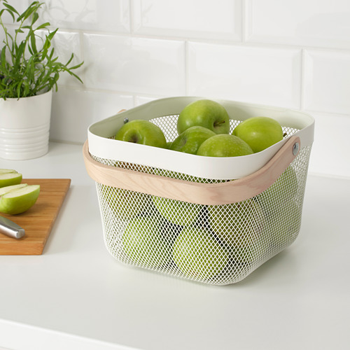 Una cesta de almacenamiento de malla con asas de madera que contiene manzanas verdes. Ventila para mantener los productos frescos. En una encimera blanca cerca de una tabla de cortar.