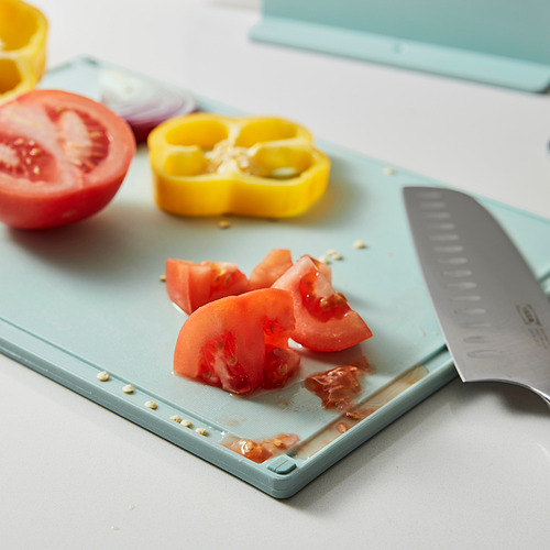 Tabla de cortar azul claro con cuchillo y trozos de tomate de colores, mostrando extremos antideslizantes y ranura para jugo.