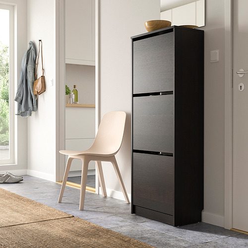 Zapatero negro con 3 compartimentos junto a una silla beige y un mueble de pared estrecho con chaquetas.