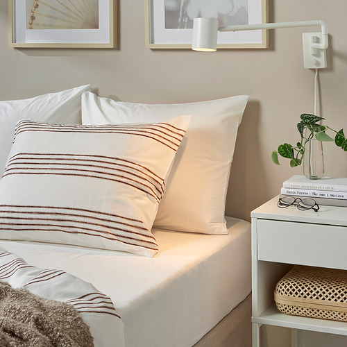 Acogedor dormitorio con ropa de cama blanca, almohada beige, libros, planta en elegante mesita de noche.