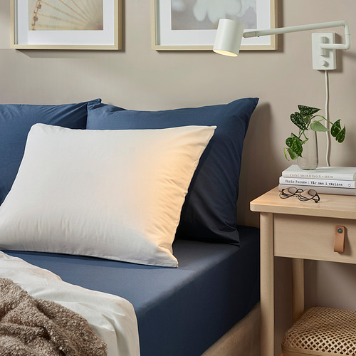 Cama con sábanas y almohadas azules ULLVIDE; Lámpara de pared blanca, libros, planta en pequeña mesita de noche de madera.