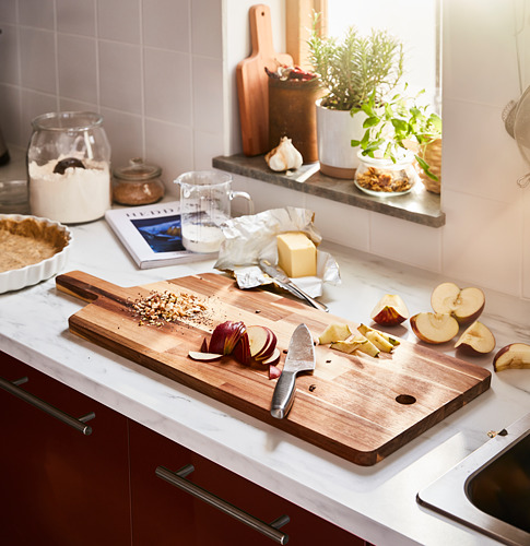 Una tabla de cortar de madera con cuchillo y rodajas de manzana se encuentra sobre una encimera de cocina blanca, rodeada de ingredientes para hornear.