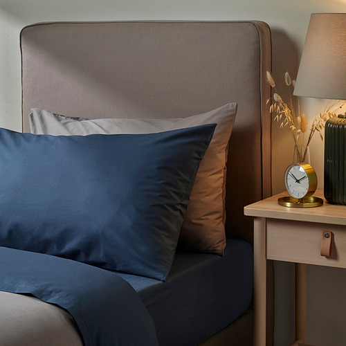 Cama con almohada ULLVIDE azul, cabecero texturizado, reloj de noche y flores secas.