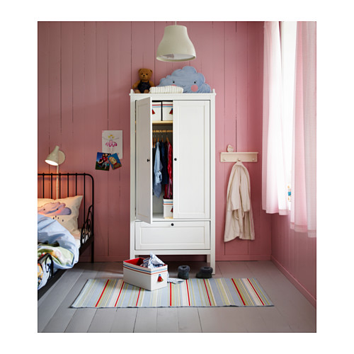 Habitación infantil con armario SUNDVIK blanco, oso, ropa, alfombra, paredes rosas, cama negra, lámpara, pósters y cortina.