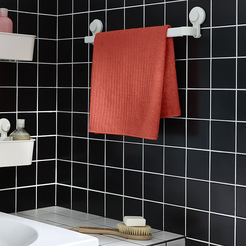 La toalla roja VÅGSJÖN cuelga entre ganchos blancos sobre una pared de azulejos oscuros. Pequeño, rectangular, adyacente al lavabo con artículos de tocador.
