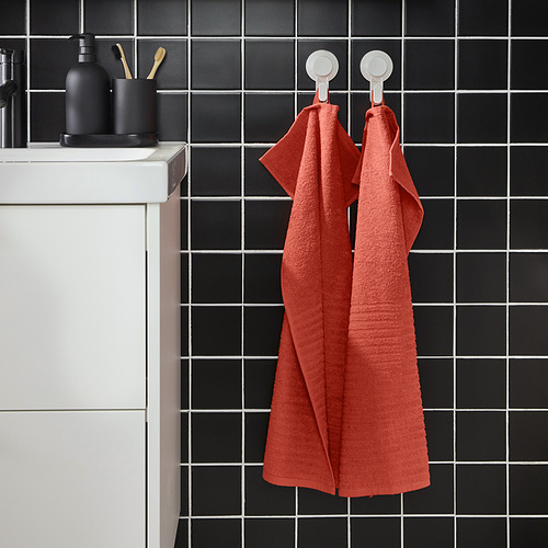 Dos vibrantes toallas VÅGSJÖN naranjas cuelgan de ganchos cerca de un fregadero, contrastando con la pared de azulejos oscuros, resaltando su tamaño y textura.