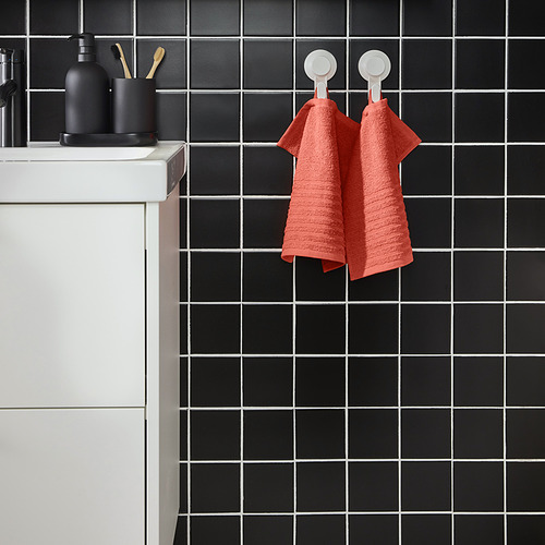 La toalla VÅGSJÖN de color naranja brillante cuelga sobre baldosas de rejilla negra, cerca del lavabo con artículos de tocador.
