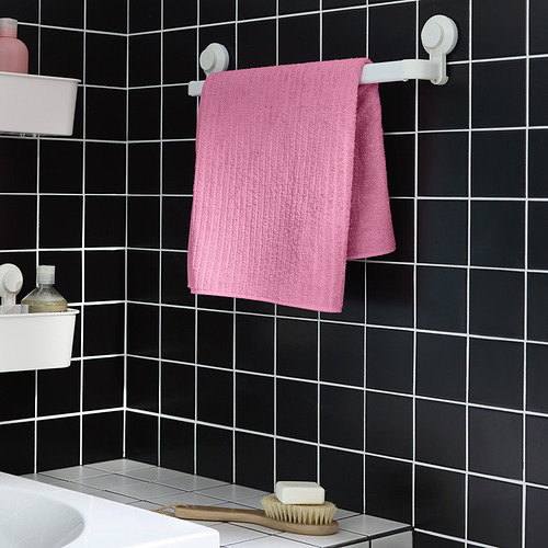 La toalla VÅGSJÖN de color rosa brillante cuelga en un soporte de pared, más pequeña que la botella adyacente.