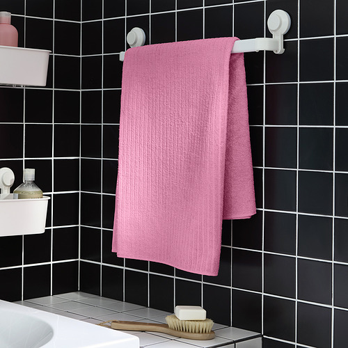 La toalla VÅGSJÖN de color rosa brillante cuelga en un estante de pared, pared de azulejos negros en contraste, con fregadero y cepillo debajo.