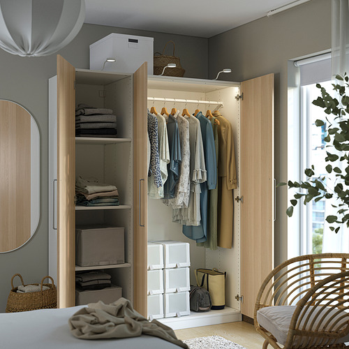 Armario moderno con puertas abiertas que muestran ropa, ropa doblada y cajas de almacenamiento. Se encuentra en una habitación luminosa con un espejo y una silla de mimbre cerca.