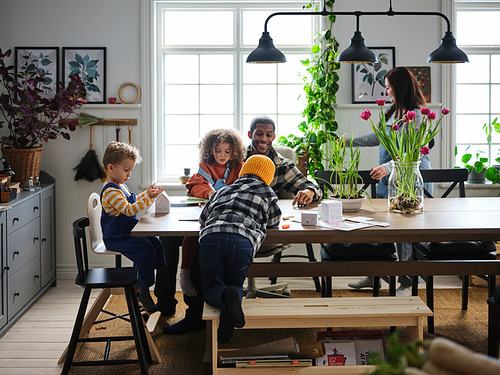 Feliz familia haciendo manualidades juntos en una acogedora mesa de madera con banco, rodeada de plantas y grandes ventanales.