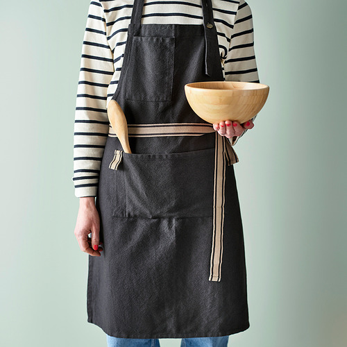 Mujer con un delantal oscuro con una camisa a rayas encima, sosteniendo un cuenco de madera. El delantal tiene bolsillos y un lazo para colgar utensilios de cocina.