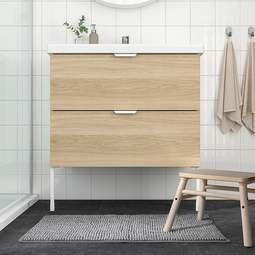 Baño moderno con tocador de madera, ducha de vidrio, alfombra gris, taburete beige y toallas