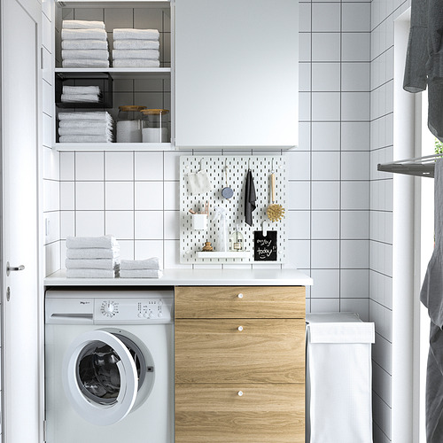 Zona de lavandería compacta con máquina de carga frontal, armario de madera, tablero perforado SKÅDIS y pared de azulejos blancos.