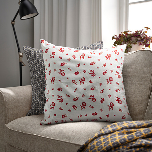 Cojín floral gris sobre sofá beige con otras almohadas, lámpara, planta.