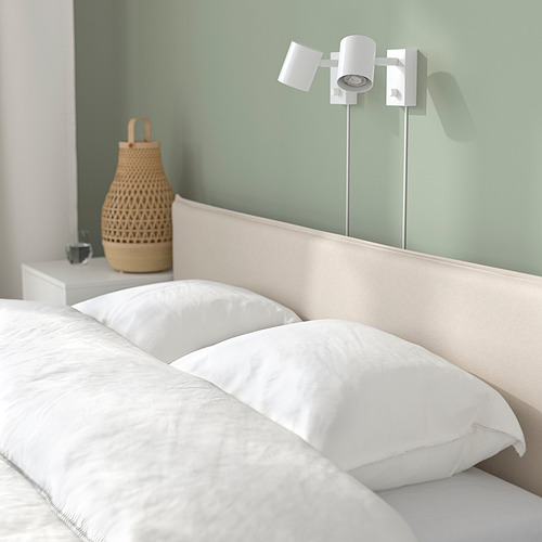 Cama minimalista con cabecero tapizado, lámparas ajustables de pared y cesta de mimbre sobre mesita de noche.