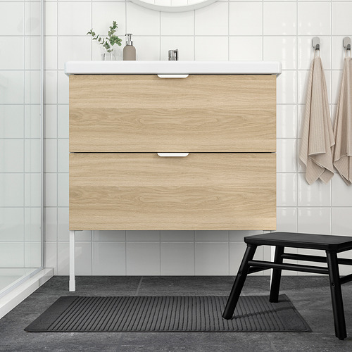 Baño moderno con tocador de madera clara, taburete negro, tapete gris, paredes blancas y puerta de vidrio.