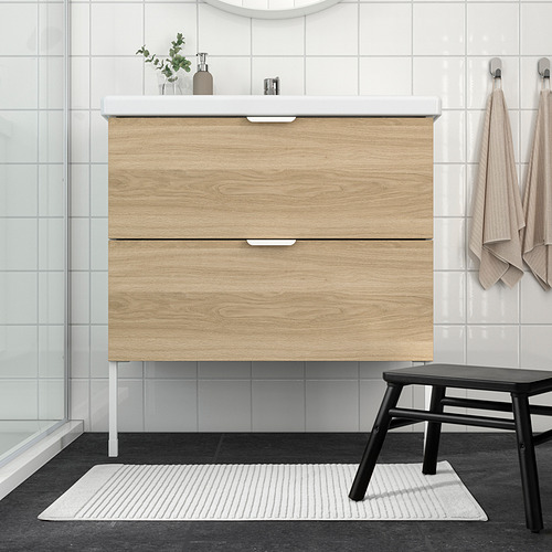 Baño moderno con tocador de madera clara, colchoneta blanca, taburete negro.