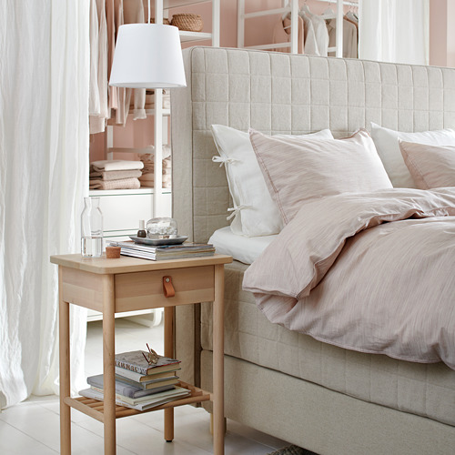 Mesita de noche BJÖRKSNÄS, lámpara, libros, cristal. Ropa de cama neutra, almohadas, cortinas transparentes. Diseño escandinavo moderno.