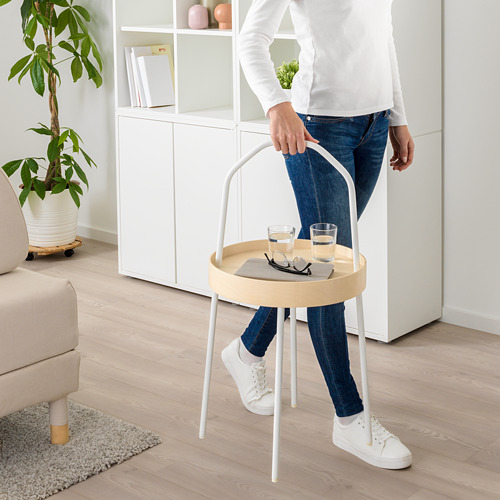 Persona que sostiene una mesa auxiliar redonda blanca con asa, que lleva dos vasos, un control remoto y un libro. La mesa es fácil de transportar por la habitación.