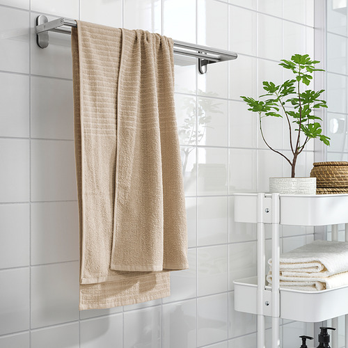 Un baño con una toalla VÅGSJÖN beige en un estante, azulejos blancos y un soporte de tres niveles con toallas y una planta.