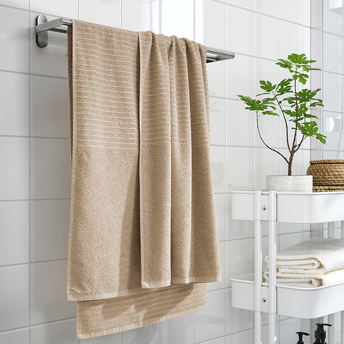 La toalla VÅGSJÖN beige cuelga en un estante de pared, cerca de una pequeña planta y toallas dobladas en un estante blanco.
