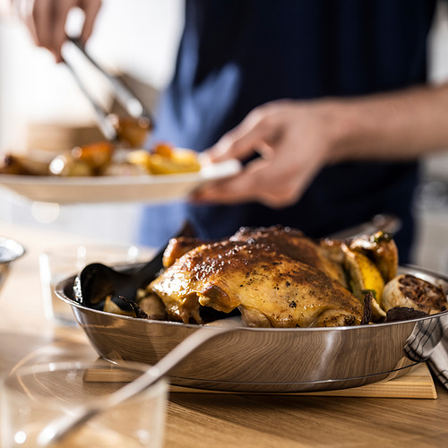 Un pollo asado en una sartén metálica se encuentra sobre una mesa de madera, listo para ser tallado con utensilios para servir.