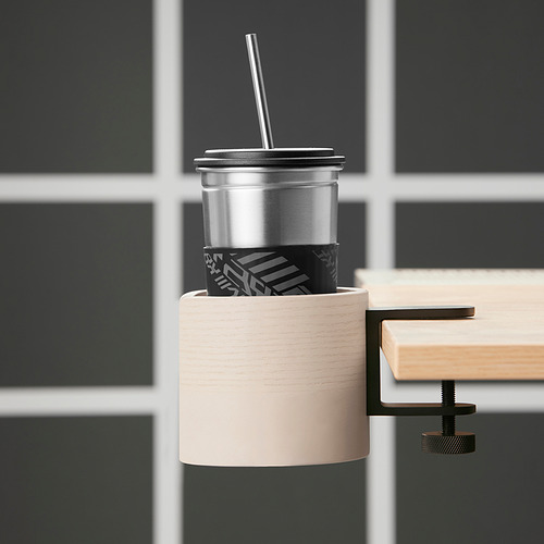 Un portatazas LÅNESPELARE de madera clara, que sujeta una taza plateada con una funda negra. El soporte está montado en un escritorio, manteniendo la bebida al alcance de la mano.