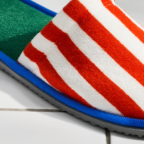 Zapatillas de rayas brillantes con suela antideslizante, con rayas naranjas y blancas, ribetes azules e interior verde sobre suelo de baldosas grises.