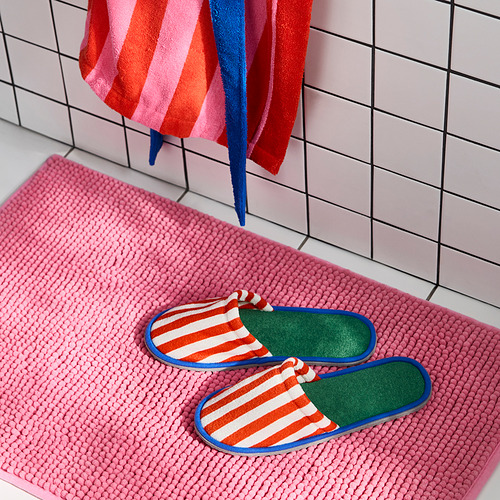Las coloridas zapatillas a rayas llamadas SKOGSSALLAT se asientan sobre una alfombra de baño rosa, más brillante que los azulejos de la pared, con una suela antideslizante para mayor estabilidad.