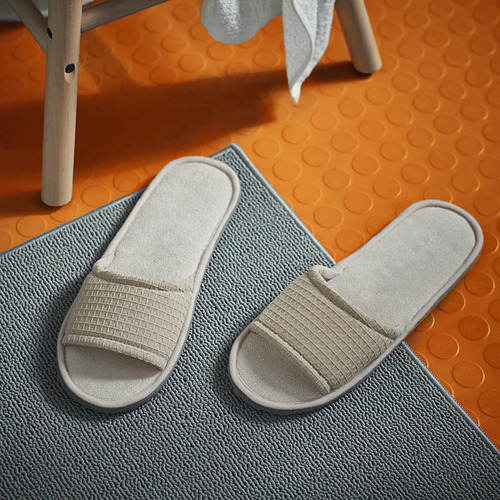 Dos zapatillas beige con textura de gofre cuidadosamente colocadas sobre una alfombra de baño azul claro. Suelas de espuma suave y diseño de punta abierta.